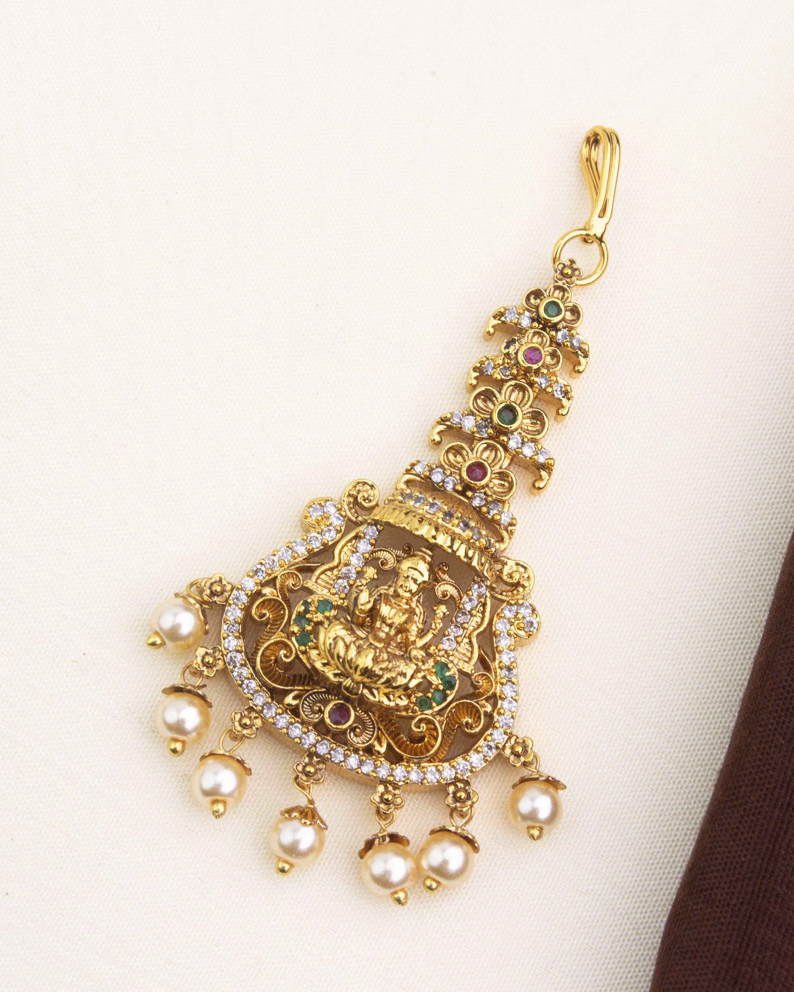 Antique gold temple maang tikka with Lakshmi motif, CZ stones, ruby and green accents, and pearl drop hangings.