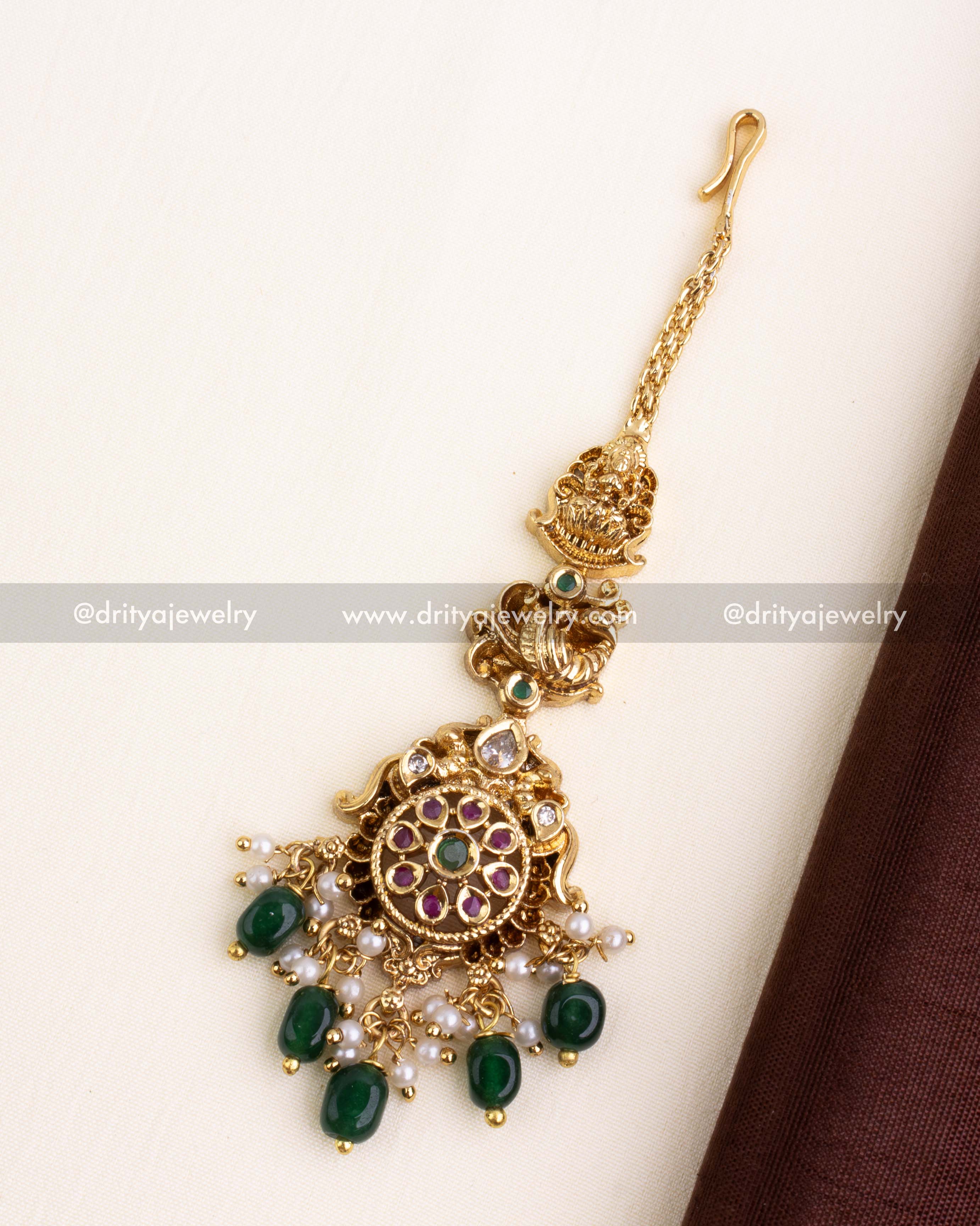 Gold-plated maang tikka with Lakshmi motif, ruby-green stones, and emerald green bead drops.