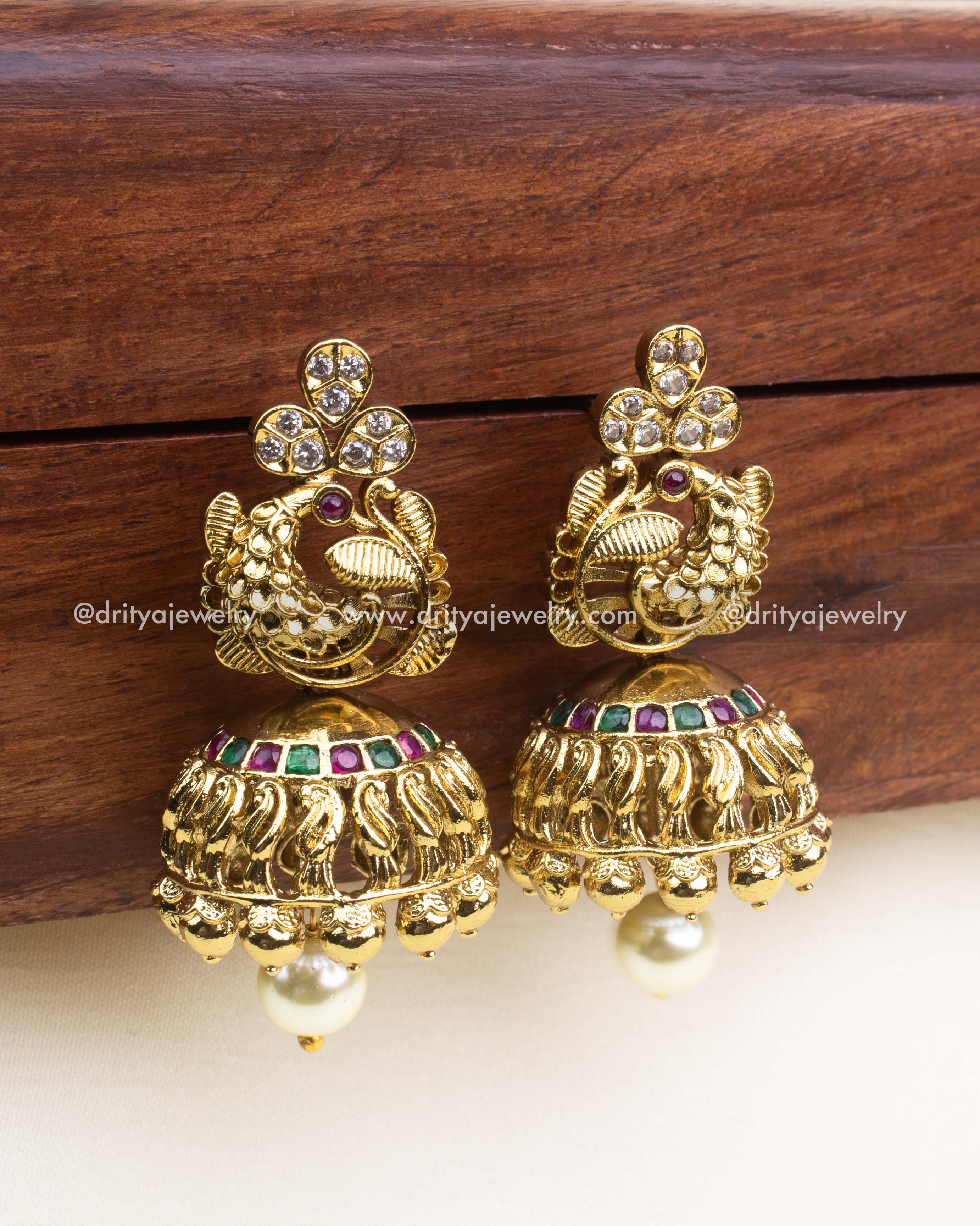 Antique gold jhumka earrings with peacock design, ruby and emerald detailing, and pearl drops.