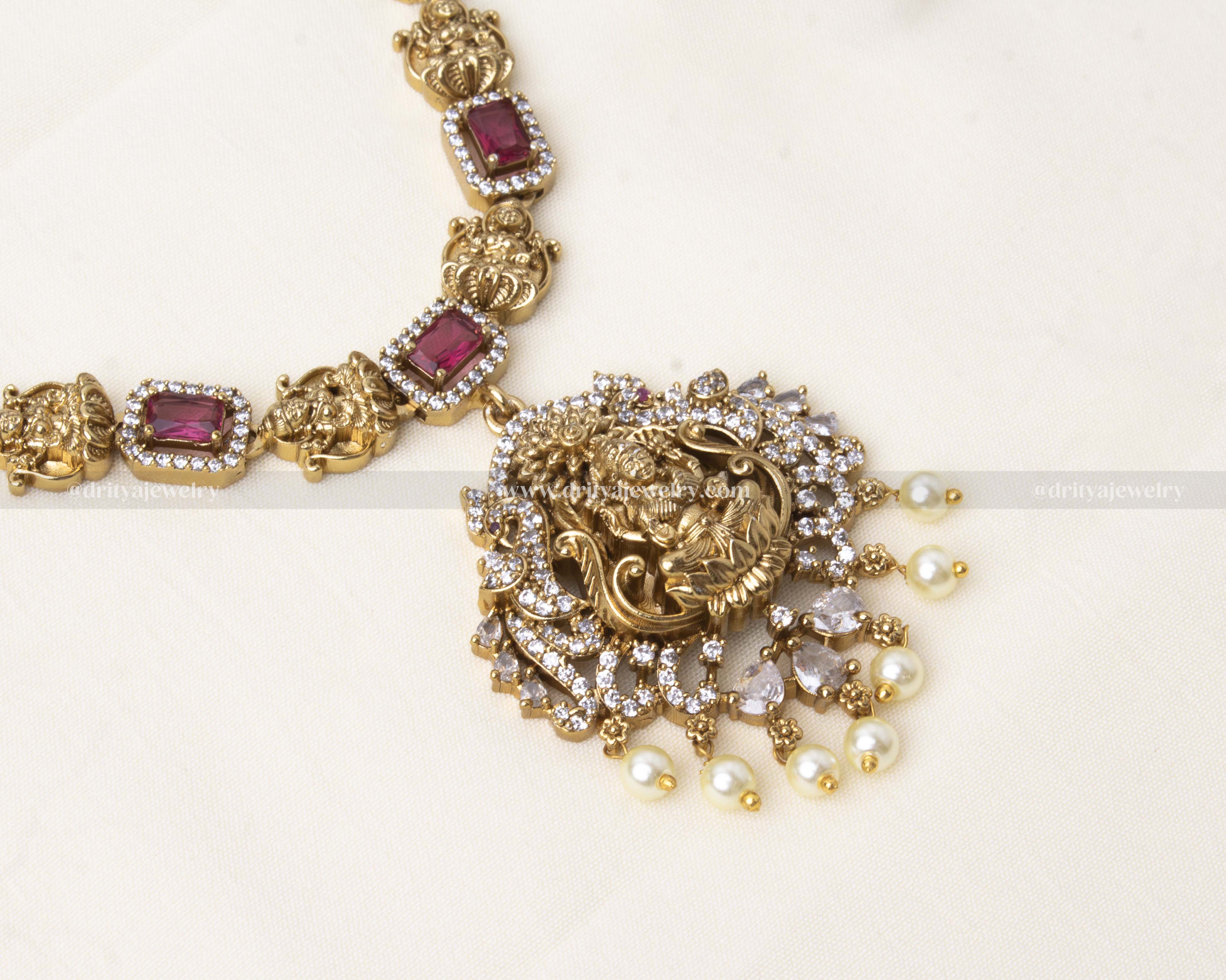 Traditional short necklace with ruby-red stones, antique gold polish, Maha Lakshmi Devi pendant, and pearl detailing, paired with matching earrings.