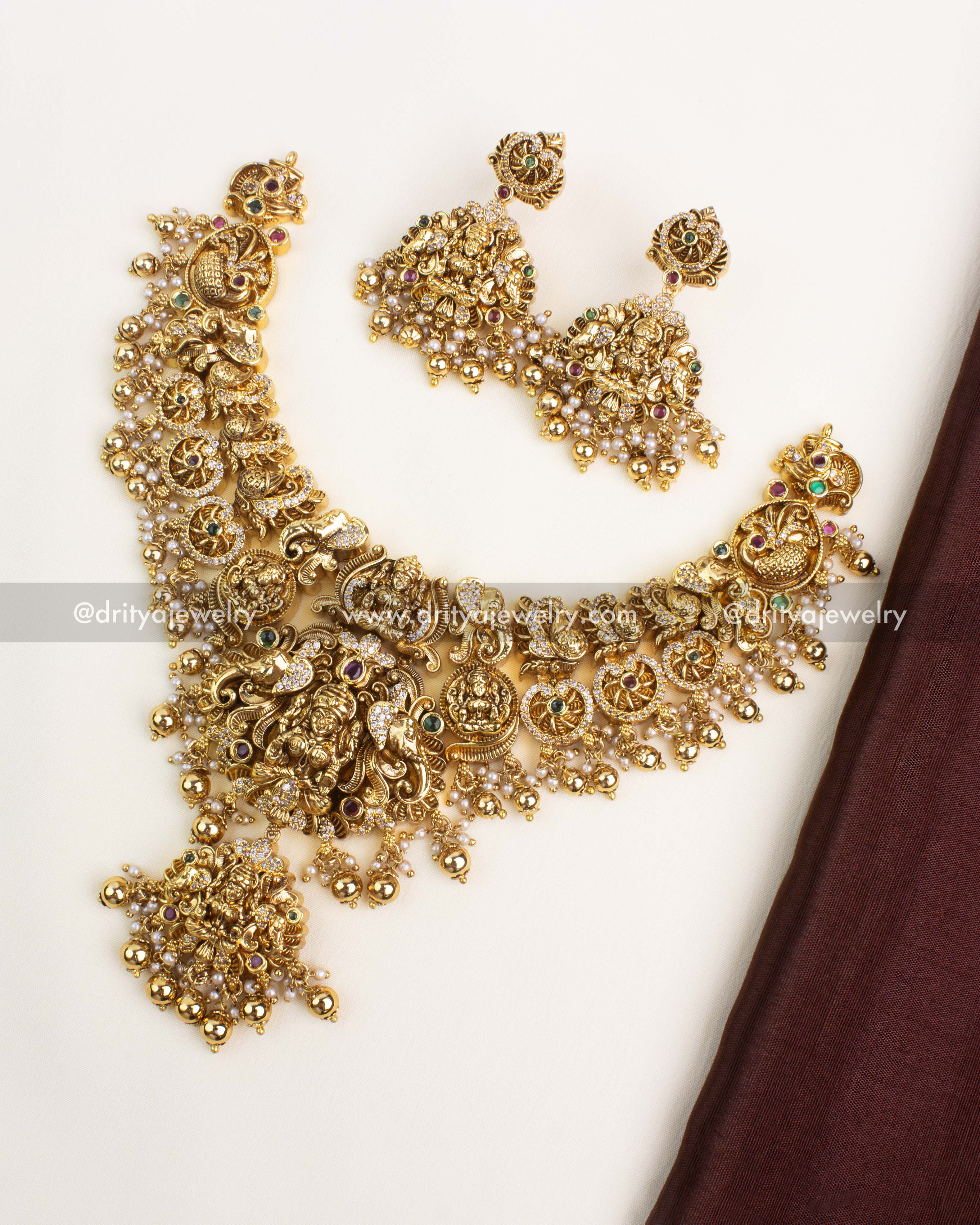 Antique gold-plated temple necklace with Lakshmi, peacock and elephant motifs, pearl clusters, and matching jhumka earrings by Dritya Jewelry.