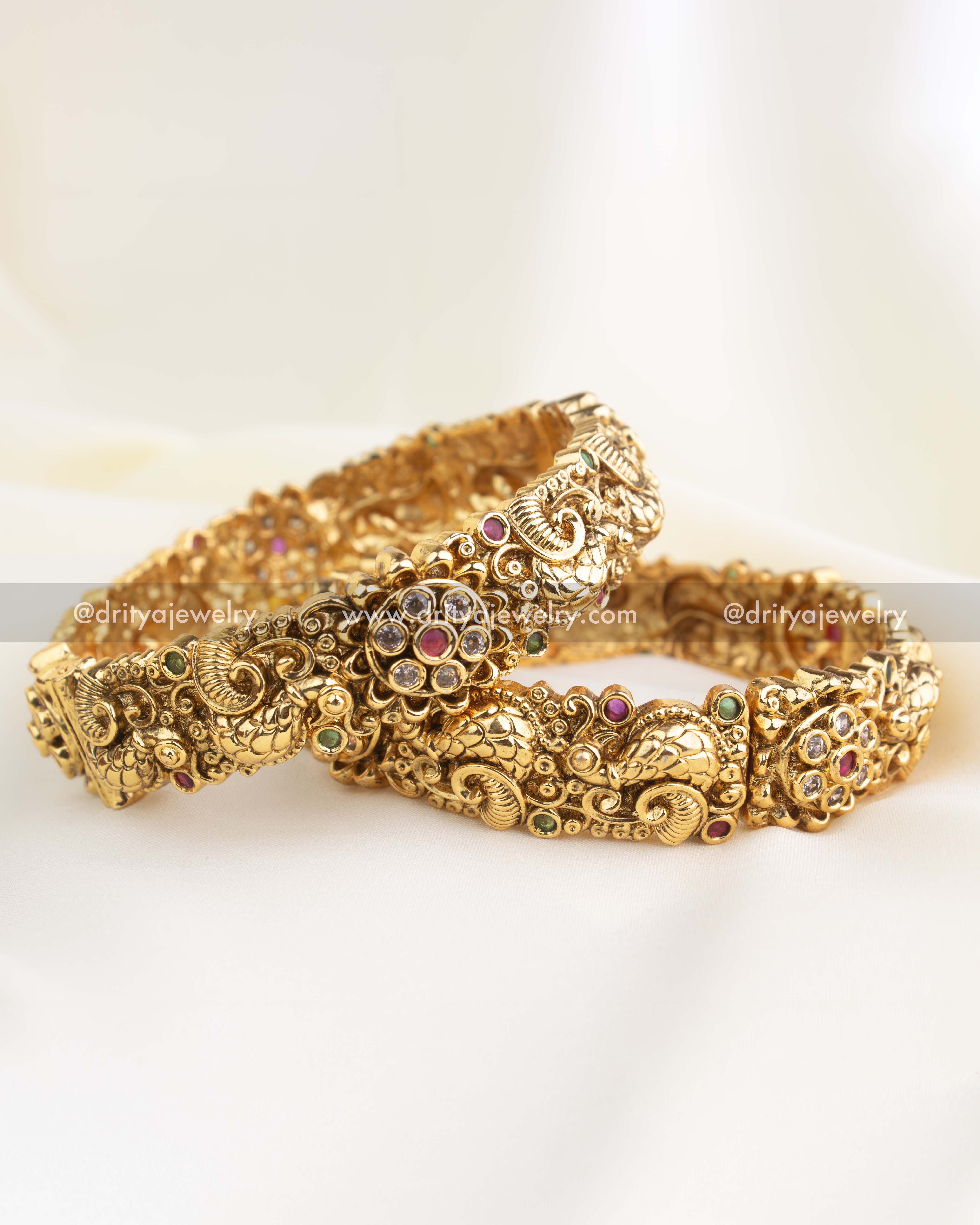 Set of antique gold Nakshi bangles with peacock carvings and CZ floral motifs in ruby and emerald tones.