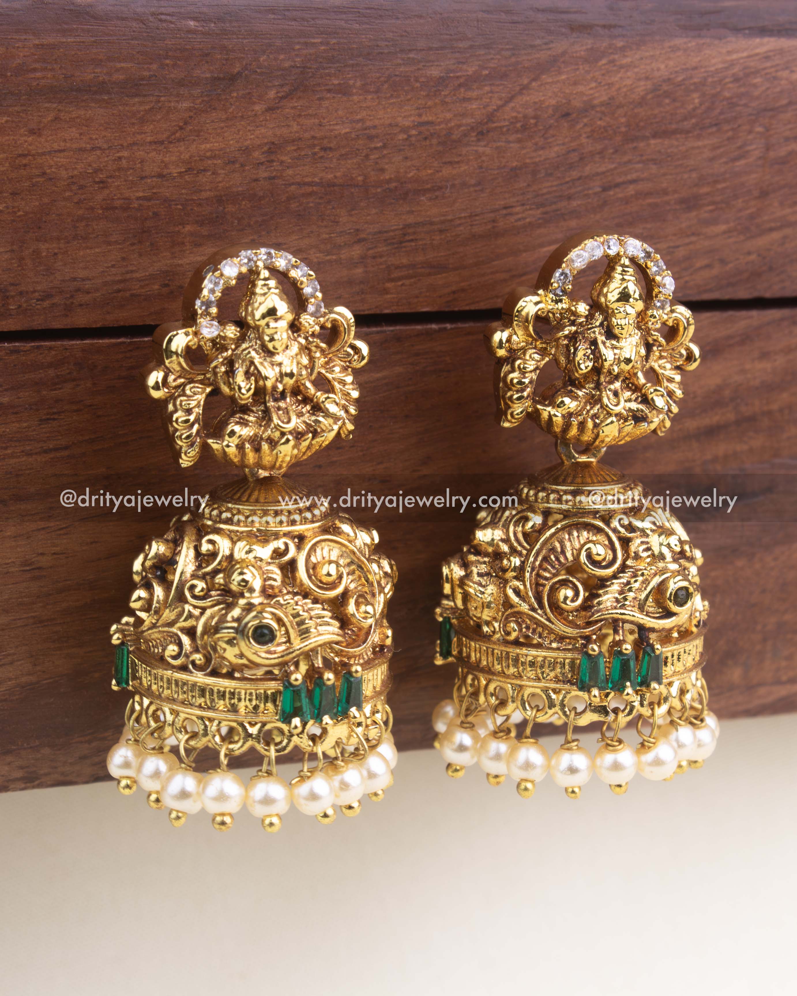 Gold-plated temple jhumka earrings with Lakshmi motif, peacock carvings, green stone accents, and pearl hangings.