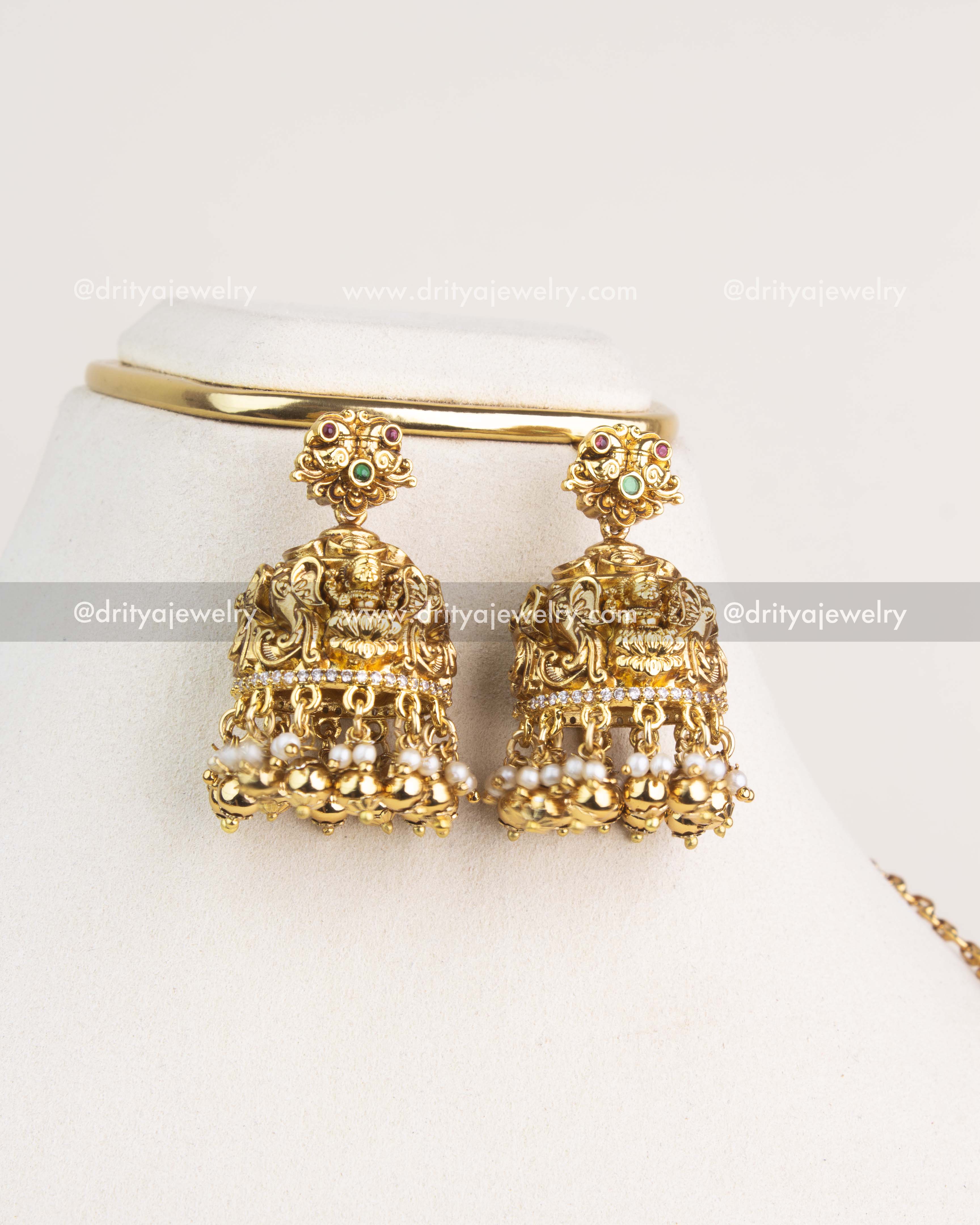 Gold-finished temple jhumka earrings featuring Lakshmi motifs, ruby and emerald stones, and pearl hangings from Dritya Jewelry.