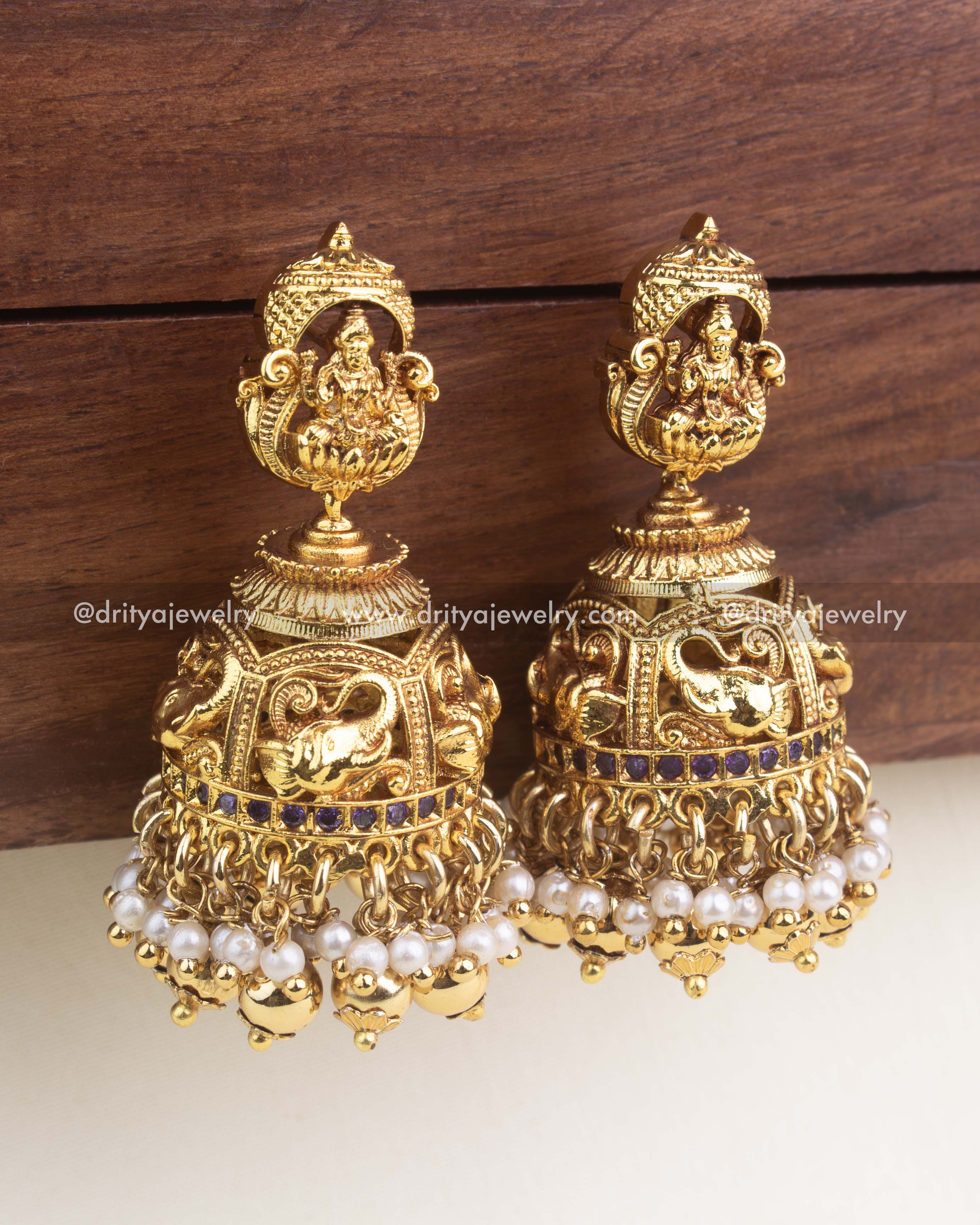 Antique gold jhumkas with Lakshmi motif, pearl clusters, and purple stone band.