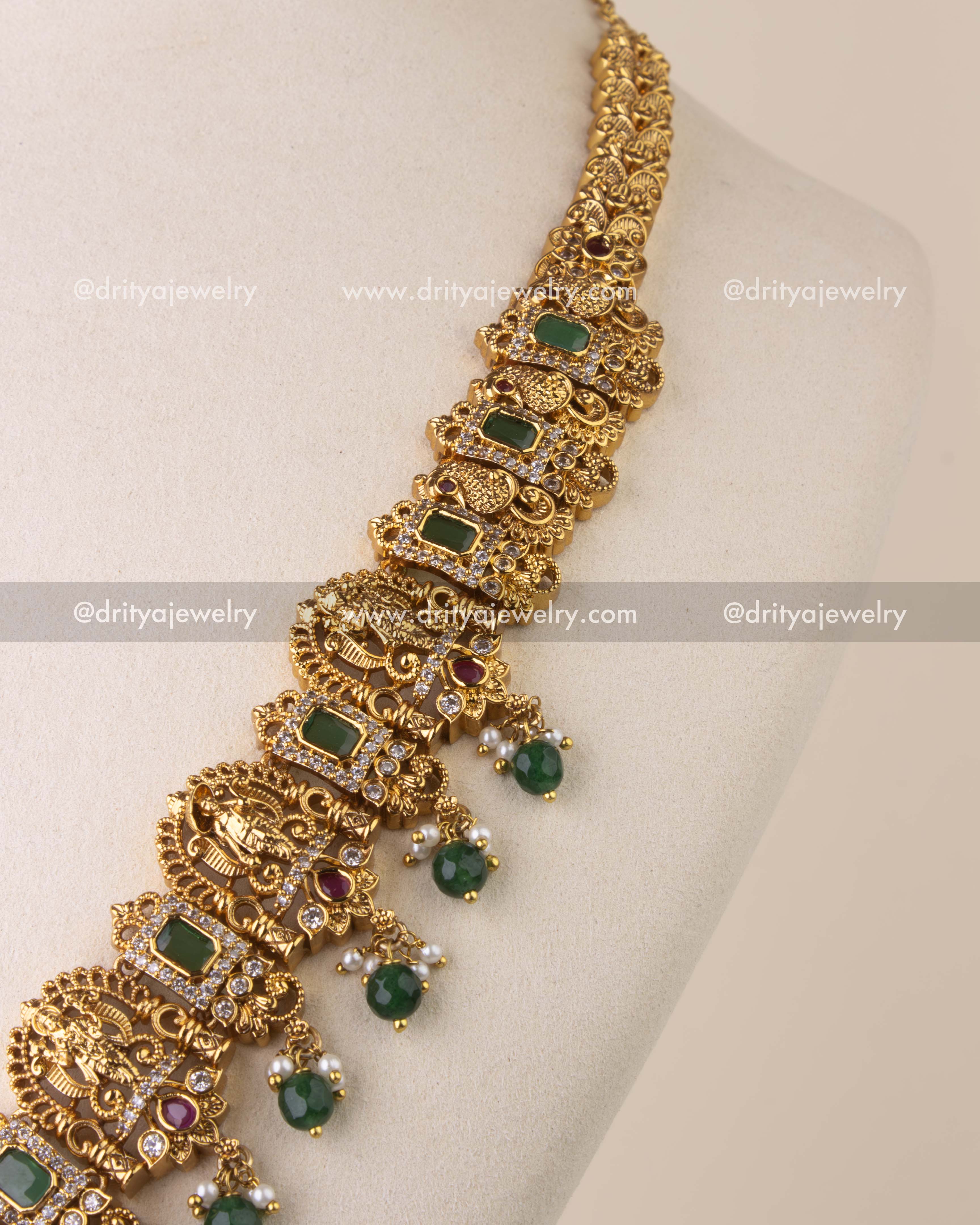 Intricate temple chain with goddess and peacock motifs, ruby embellishments, and emerald rectangular stones.