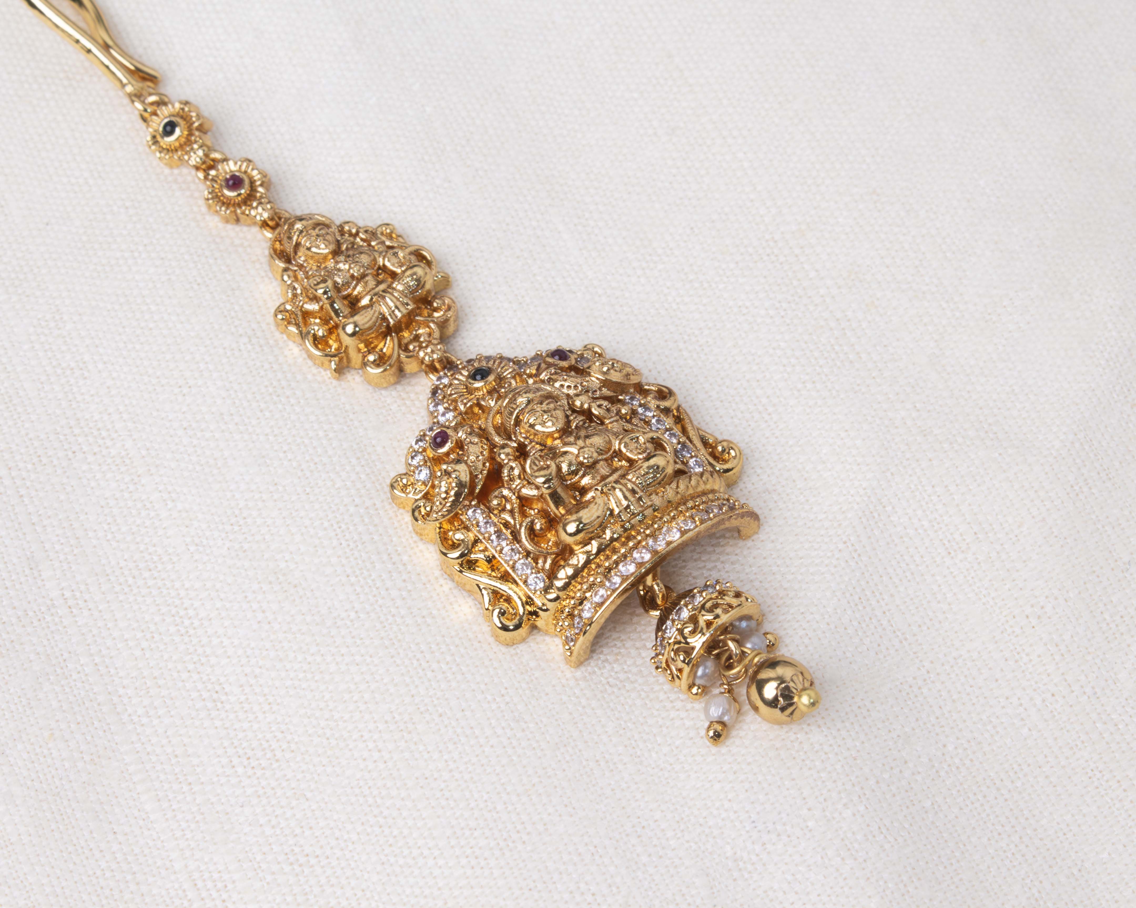 Gold polished Maang Tikka featuring a detailed Lakshmi Devi motif adorned with purple gemstones and pearl accents, ideal for spiritual and traditional occasions.