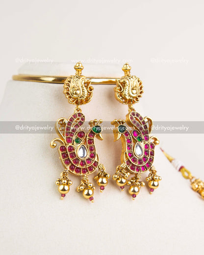 Peacock-inspired earrings with ruby stones, kundan work, and gold bead hangings in antique finish.