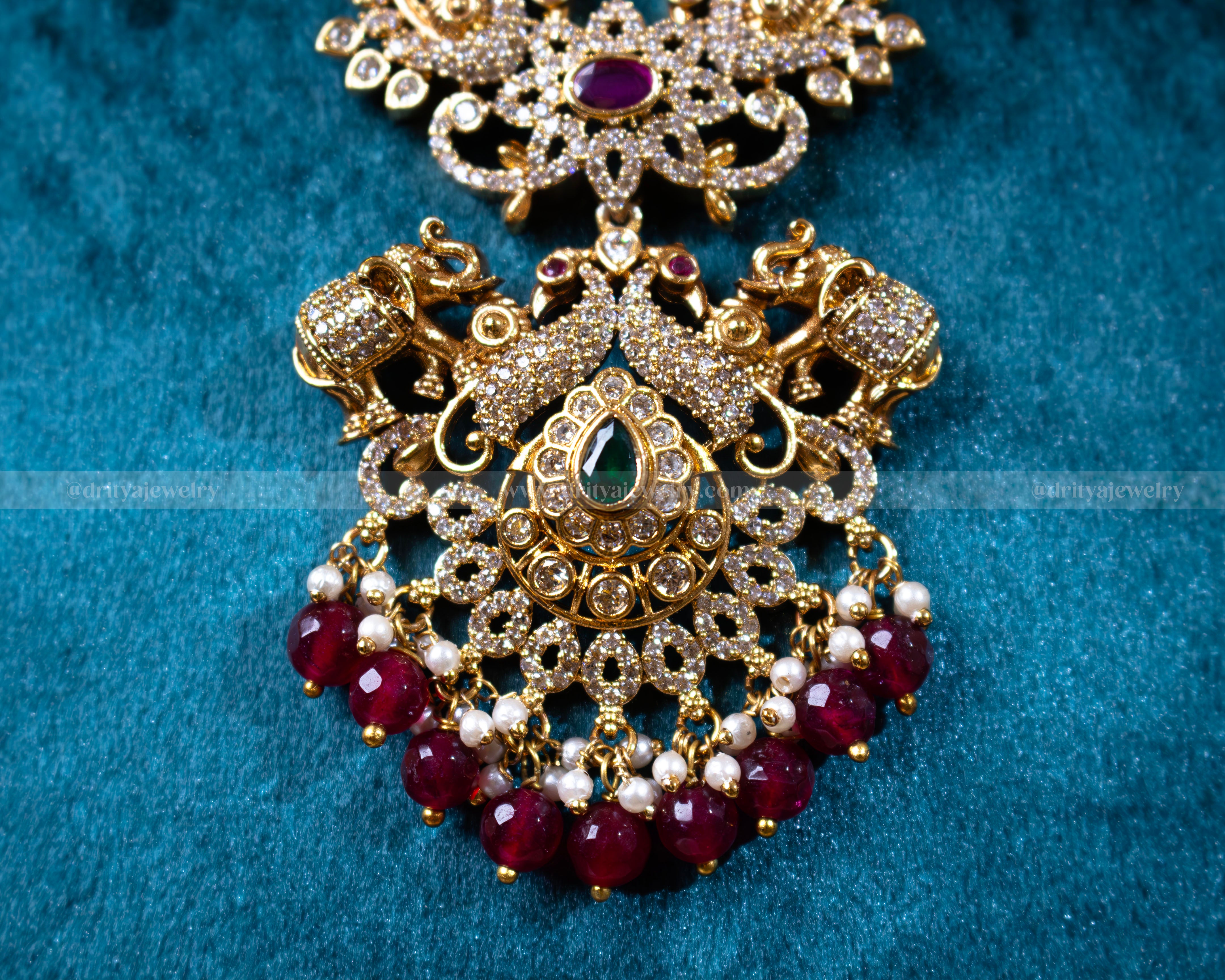 Close-up of the elephant and peacock motif pendant from Dritya Jewelry's antique gold finish necklace set, adorned with red beads and stones.