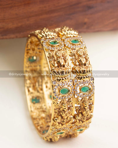 Gold temple bangles with intricate goddess motifs and oval emerald green stones surrounded by AD detailing