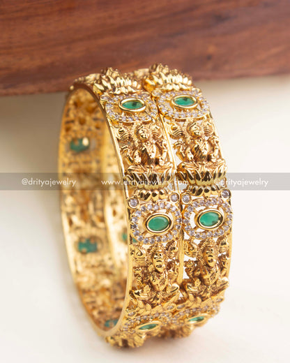 Gold temple bangles with intricate goddess motifs and oval emerald green stones surrounded by AD detailing