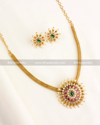 Traditional sunburst necklace with ruby-red and green stones, pearl danglers, and antique gold finish.