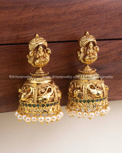 Close-up of premium temple jhumka showing Goddess Lakshmi, nakshi dome carving, green stones, and pearl embellishments.