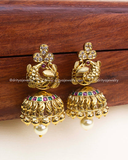 Antique gold jhumka earrings with peacock design, ruby and emerald detailing, and pearl drops.