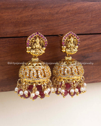 Temple-style jhumkas with Lakshmi motif, CZ detailing, and ruby–pearl drops.