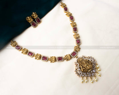 Traditional short necklace with ruby-red stones, antique gold polish, Maha Lakshmi Devi pendant, and pearl detailing, paired with matching earrings.