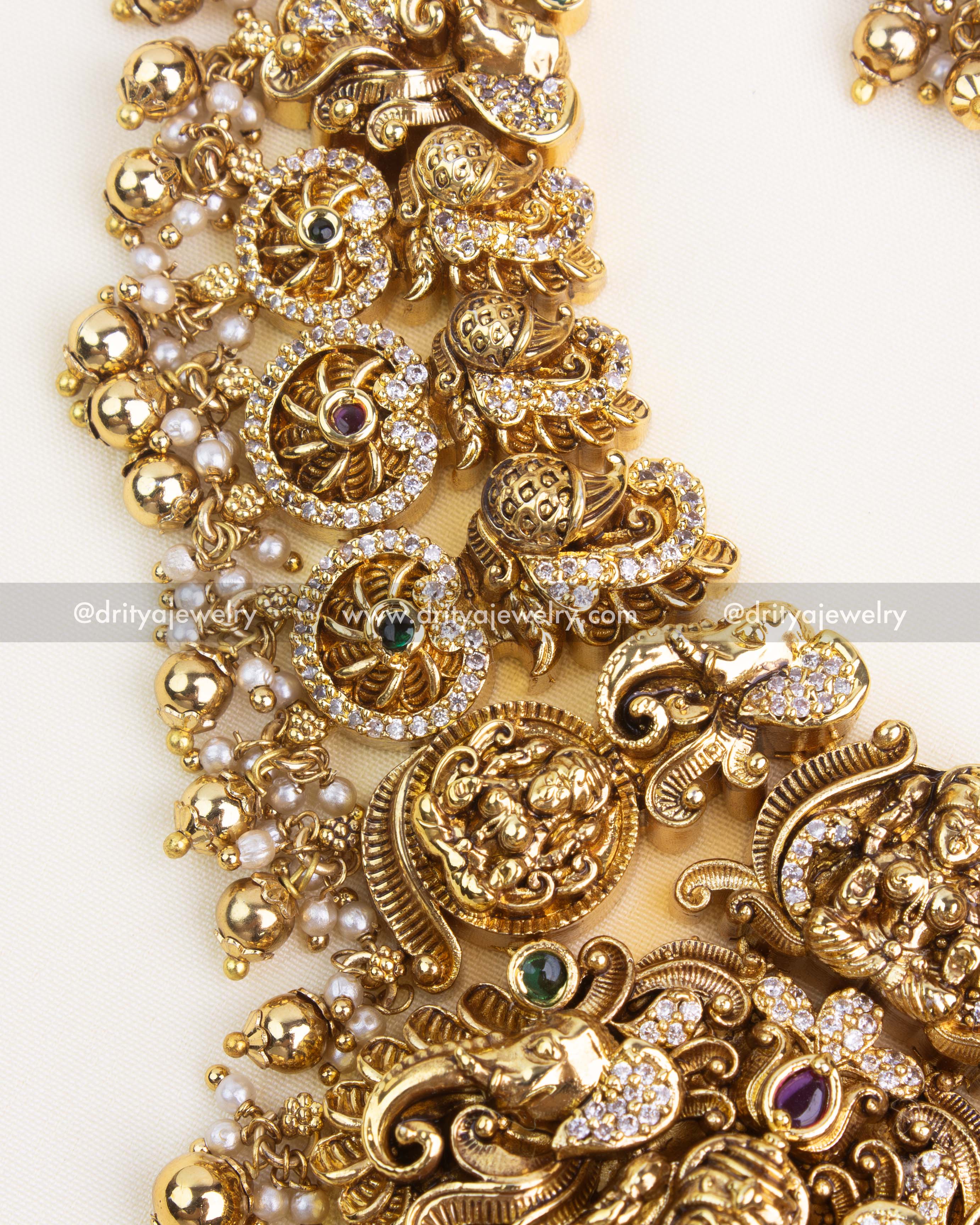 Close-up of nakshi carving with coin medallions, ruby–emerald accents, and micro pavé AD stones on antique gold finish.