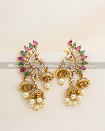Gold-tone peacock motif jhumka earrings with ruby-green stones and pearl drops.