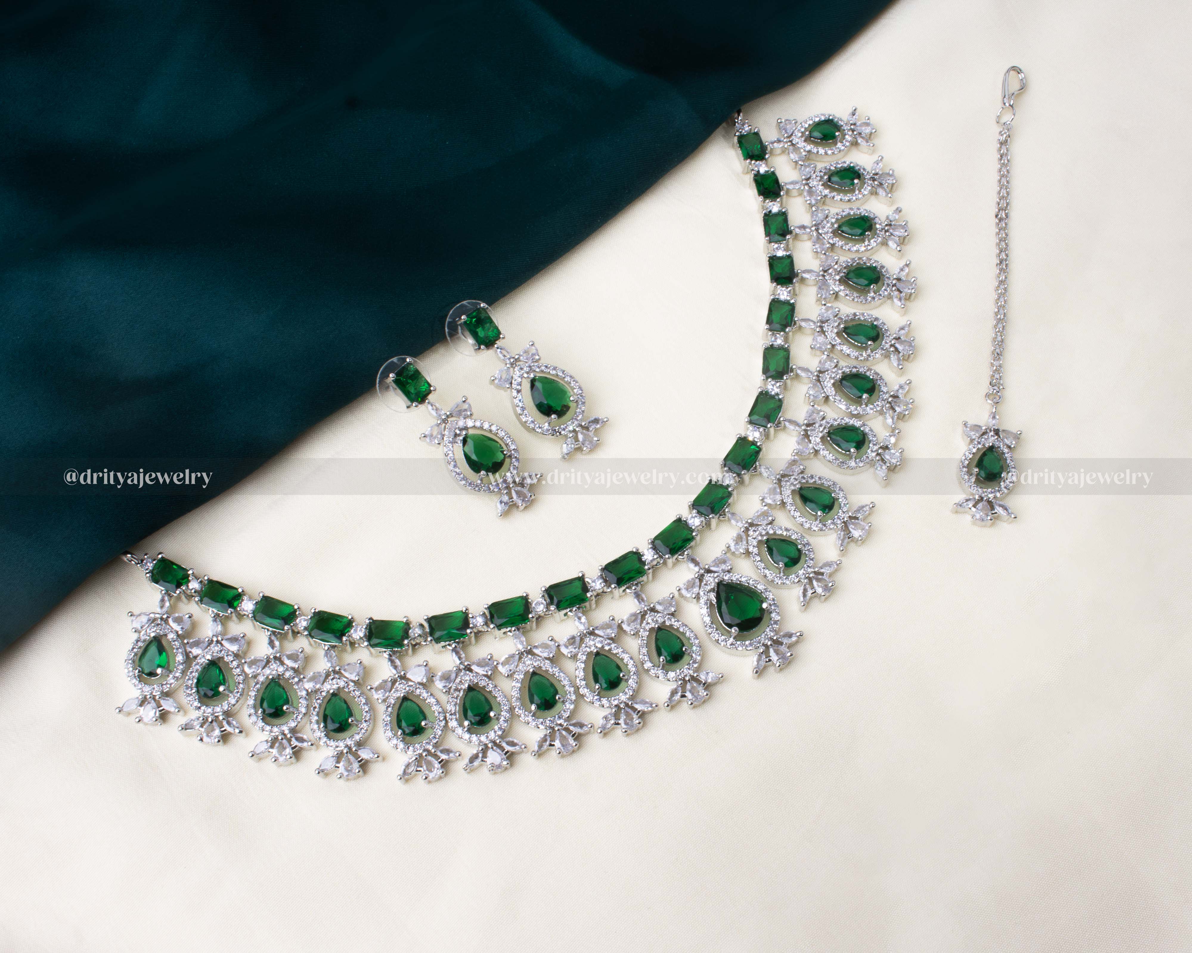 Elegant American diamond necklace set featuring green stones, matching earrings, and a maang tikka with a silver-toned finish.