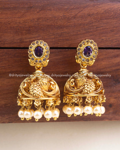 Antique gold jhumka earrings featuring peacock carvings, purple stone tops, CZ detailing, and pearl drop hangings.