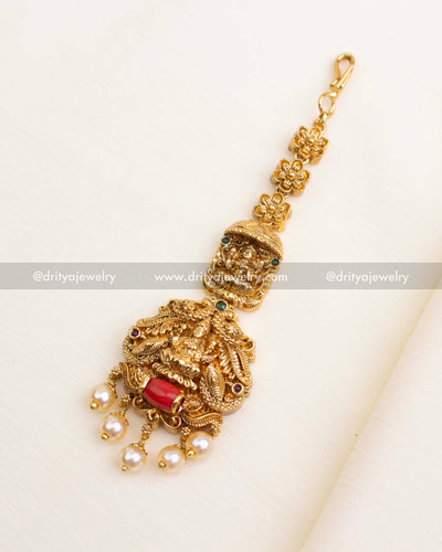 Temple-style nakshi maang tikka featuring goddess motif, peacock carvings, red coral bead, and pearl drops.