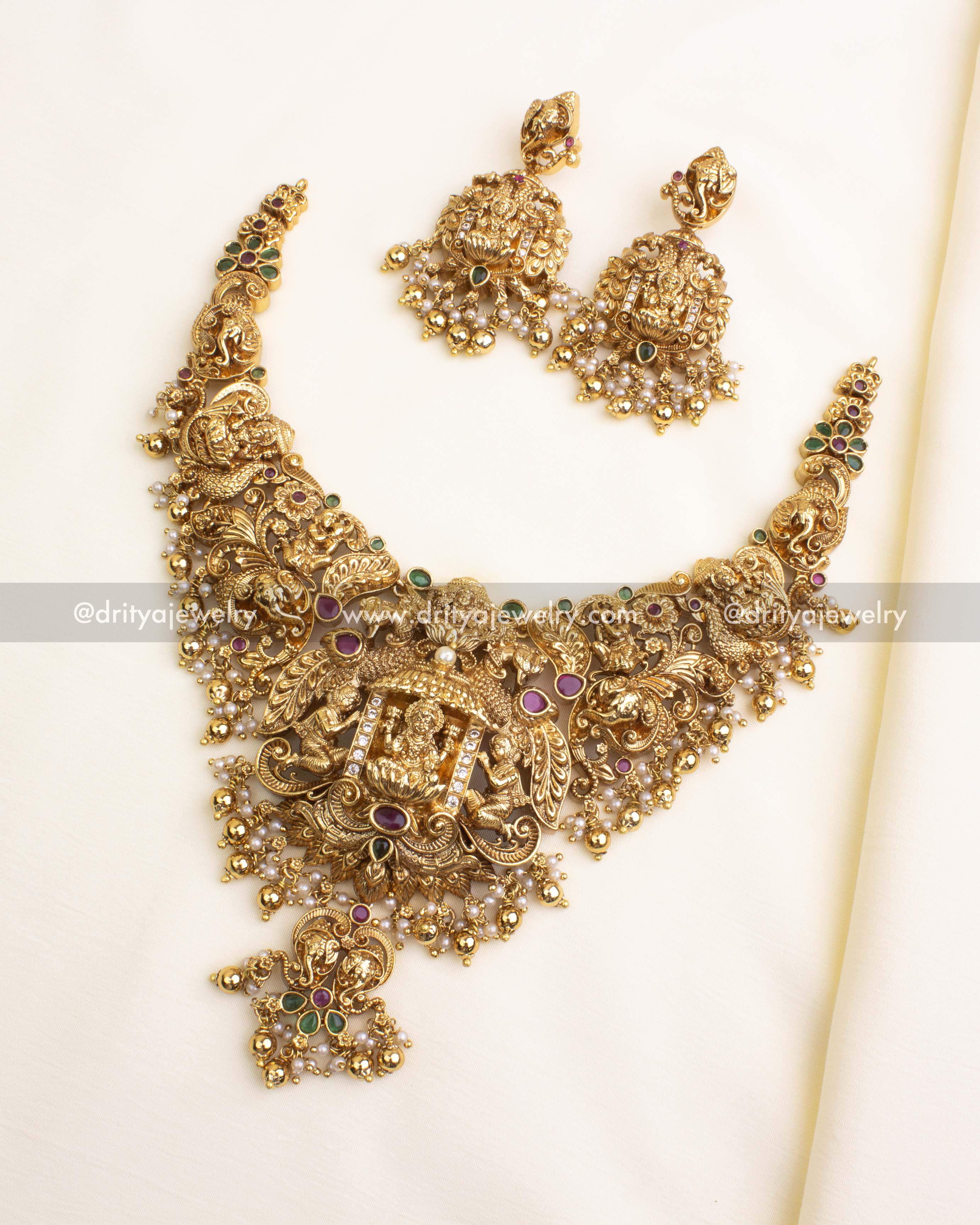 Temple-style nakshi short necklace with goddess centerpiece, peacock and elephant carvings, green and ruby stones, and pearl-gold bead drops.