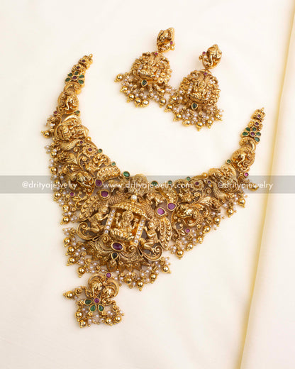 Temple-style nakshi short necklace with goddess centerpiece, peacock and elephant carvings, green and ruby stones, and pearl-gold bead drops.