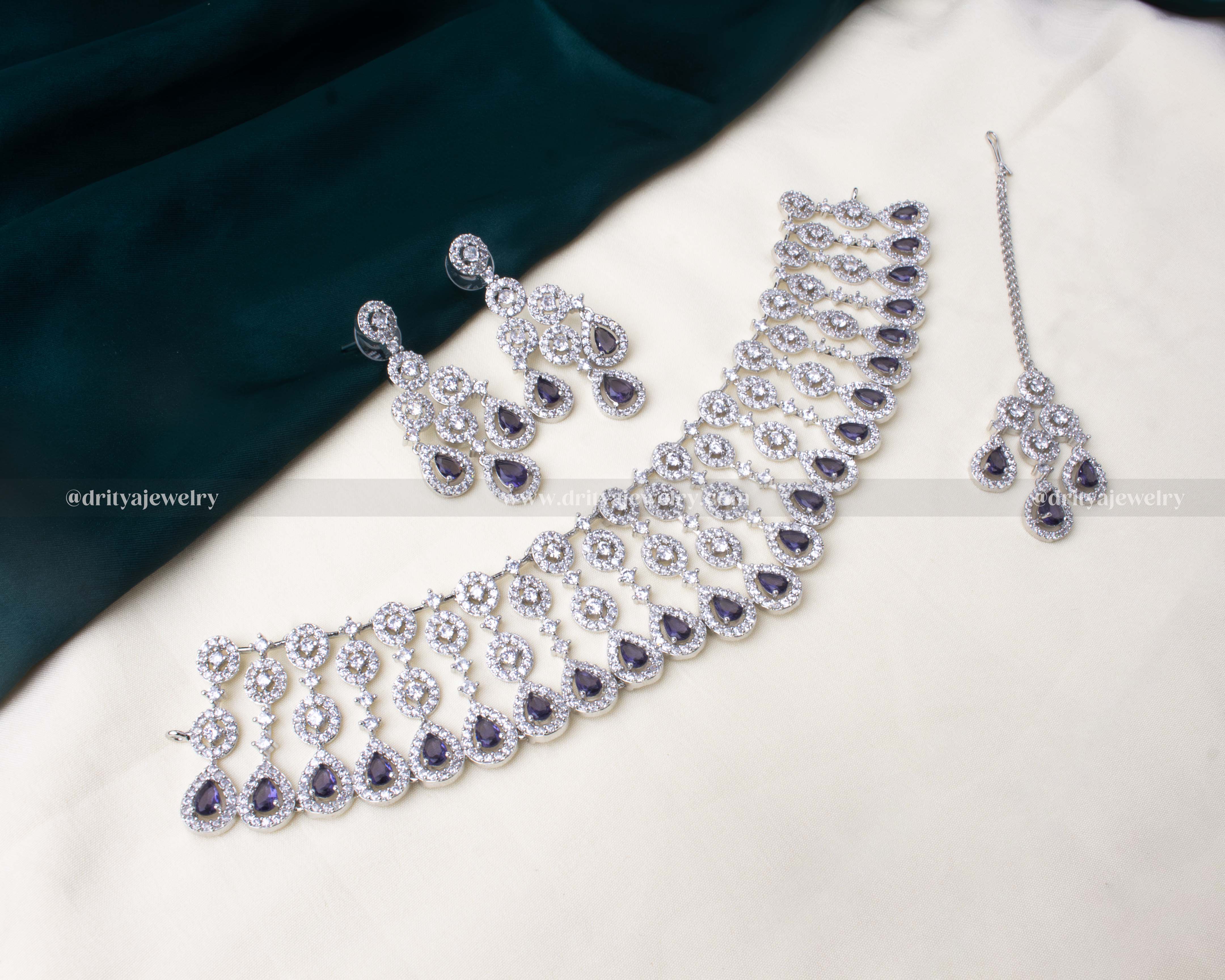 Elegant zircon choker necklace set with American diamonds, cascading teardrop design, paired with dangler earrings and a maang tikka in white and purple colors.