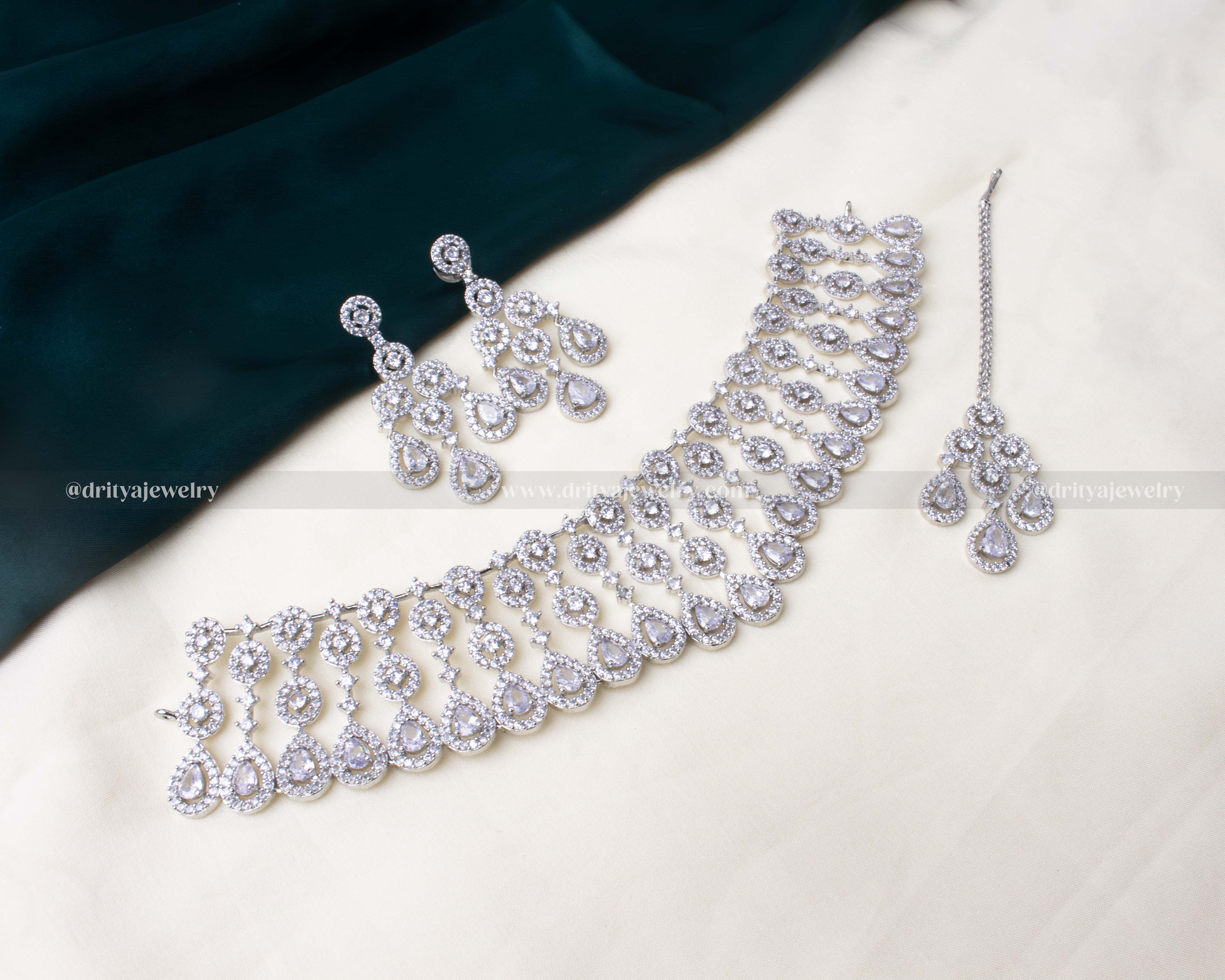 Elegant zircon choker necklace set with American diamonds, cascading teardrop design, paired with dangler earrings and a maang tikka in white and purple colors.