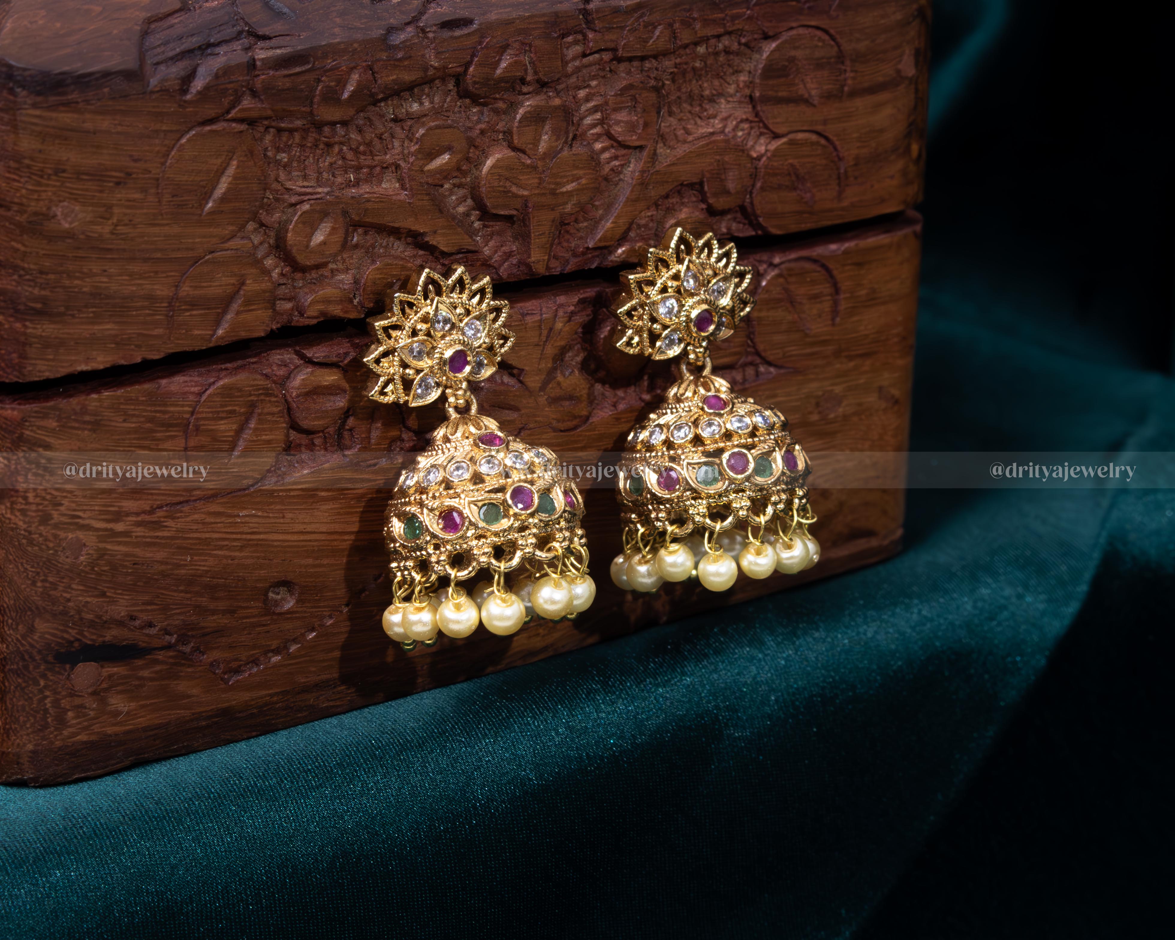 Opulent gold jhumka earrings with ruby and emerald accents and pearl details, ideal for bridal and festive wear.