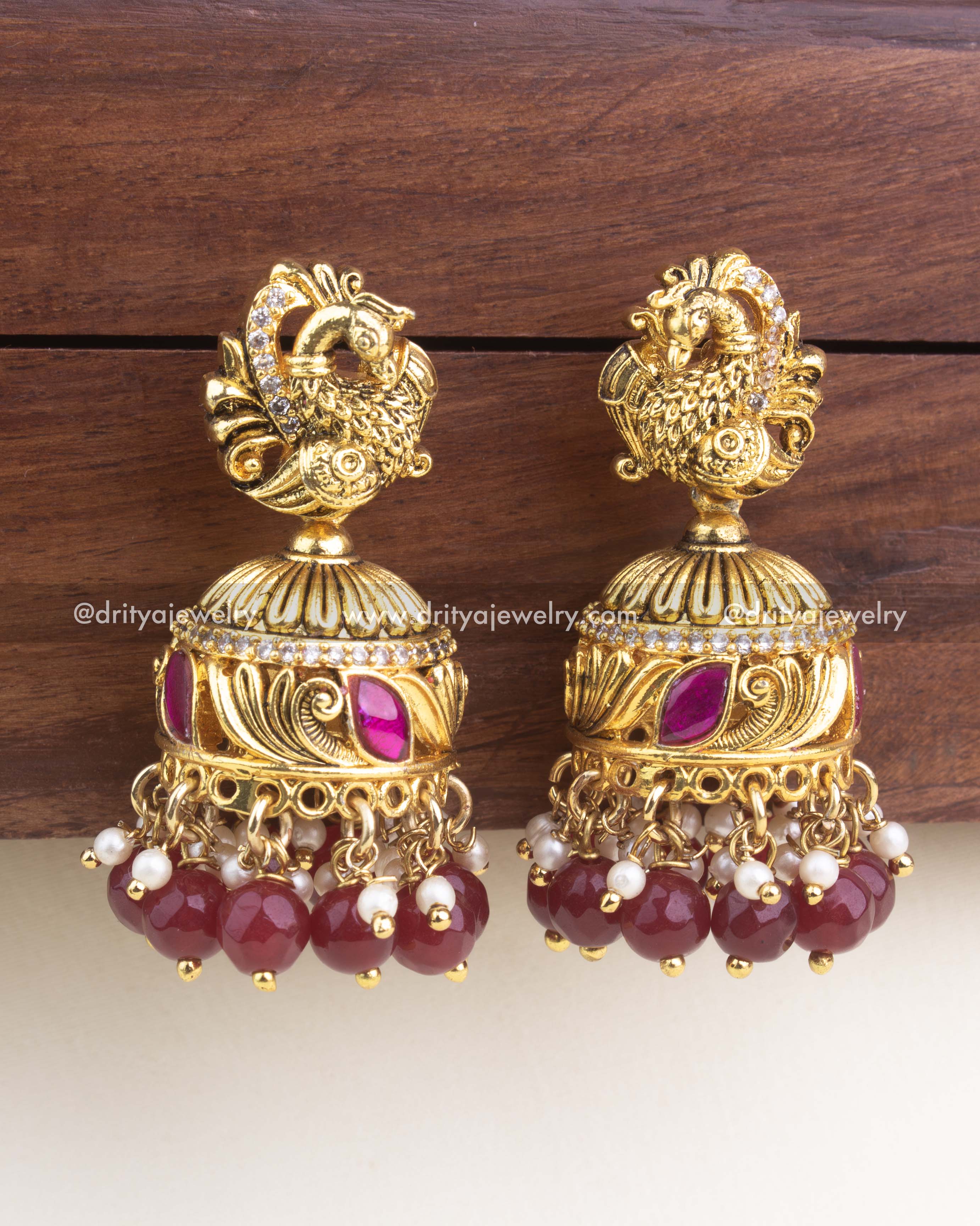 Gold-plated kemp jadau jhumka with peacock top, ruby red enamel accents, and deep ruby bead hangings.