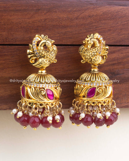Gold-plated kemp jadau jhumka with peacock top, ruby red enamel accents, and deep ruby bead hangings.