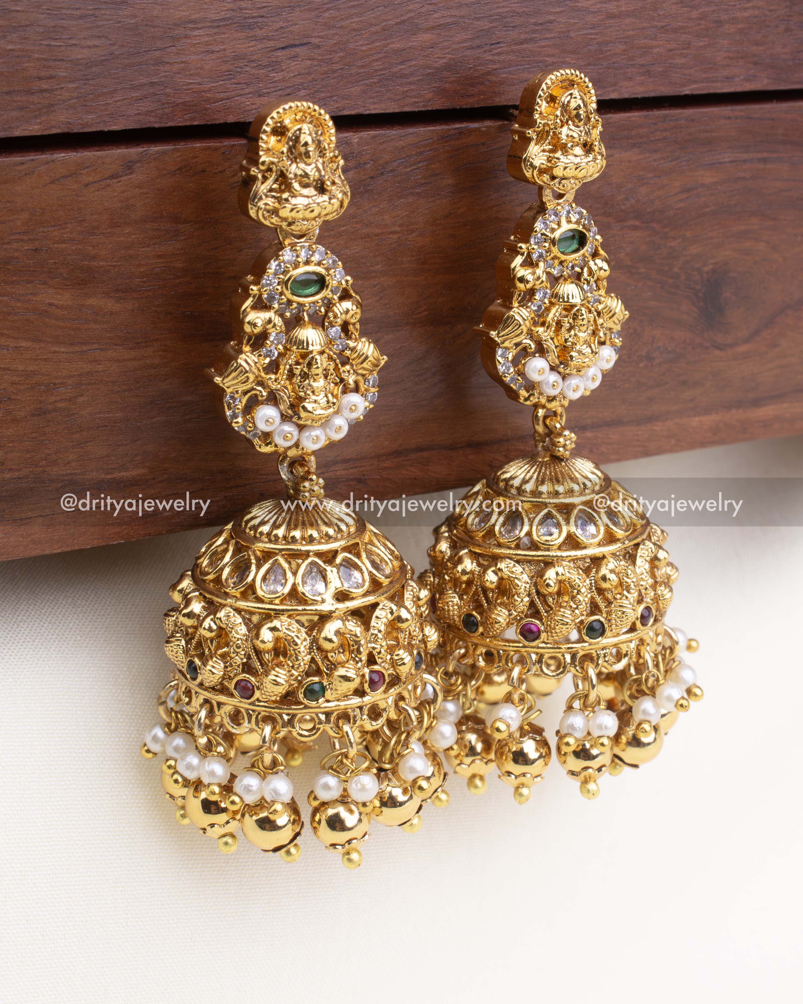 Antique-finish jhumka earrings with Lakshmi Devi design, peacock dome, and pearl-gold ball drops.