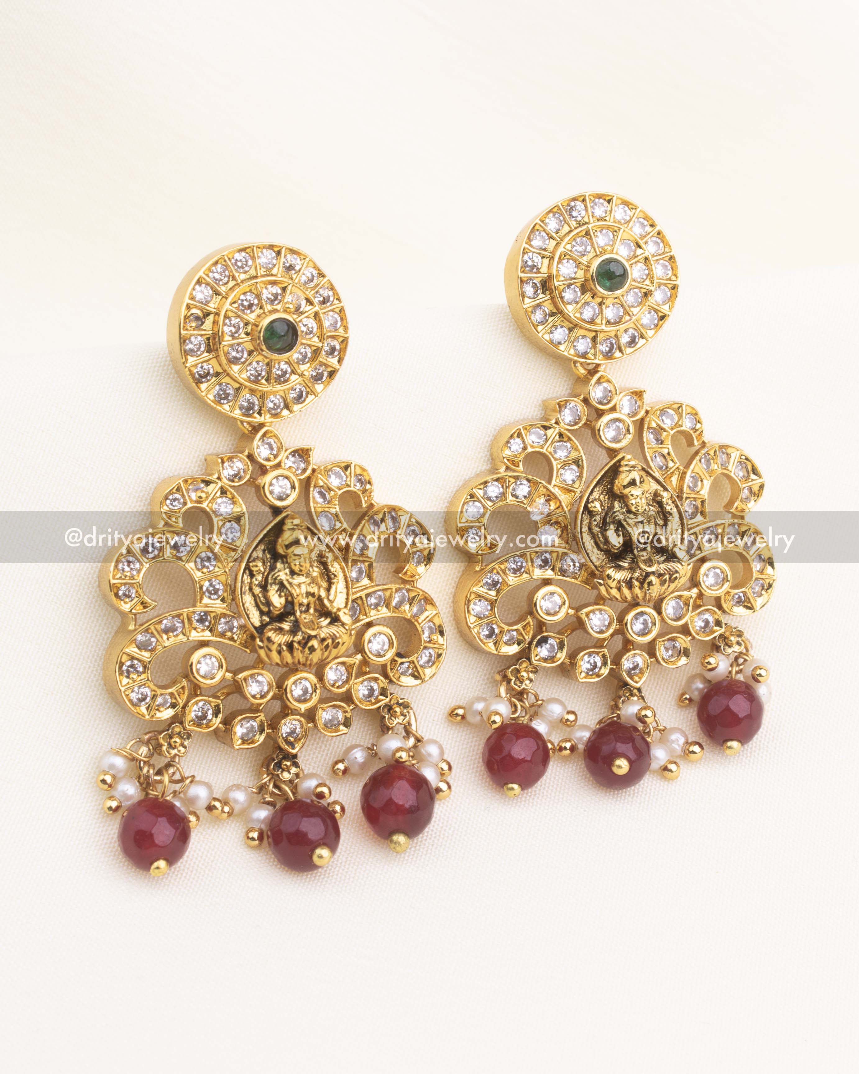 Matching temple danglers with Lakshmi panel, AD stones, and ruby red bead drops in antique gold finish.