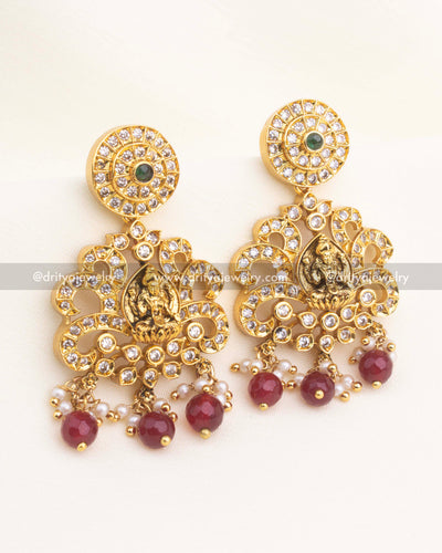 Matching temple danglers with Lakshmi panel, AD stones, and ruby red bead drops in antique gold finish.