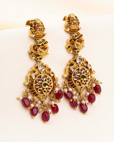 Gold temple earrings with traditional peacock and goddess motifs, finished with ruby red beads and pearl hangings.