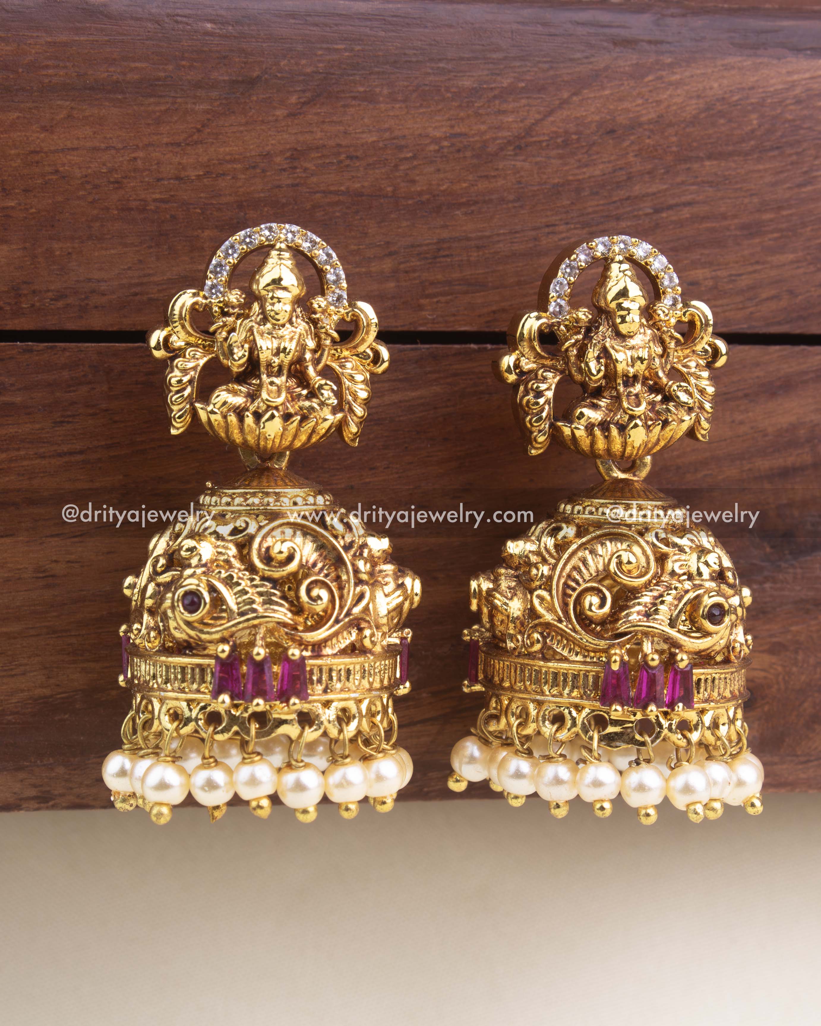 Premium temple-style jhumka earrings with Lakshmi motif, red stone accents, peacock carvings, and pearl hangings.