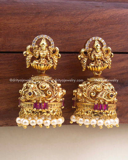Premium temple-style jhumka earrings with Lakshmi motif, red stone accents, peacock carvings, and pearl hangings.