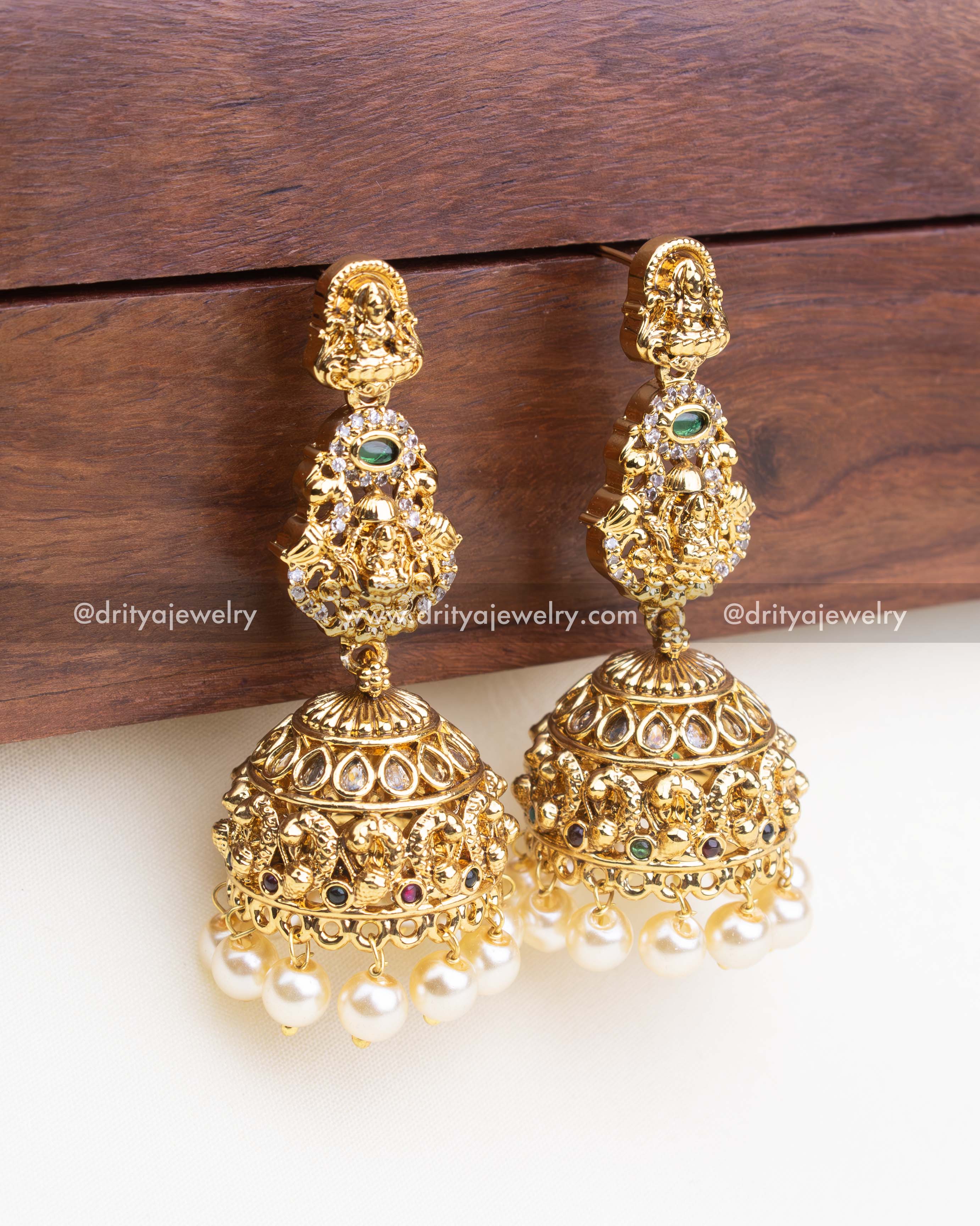 Pair of dome-shaped temple jhumkas with Lakshmi Devi carvings, ruby-emerald accents, and traditional pearl fringe.
