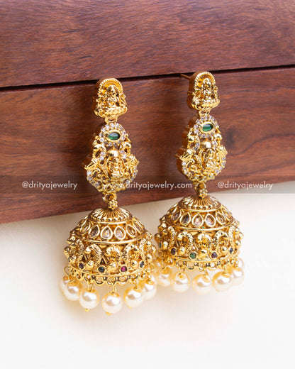 Pair of dome-shaped temple jhumkas with Lakshmi Devi carvings, ruby-emerald accents, and traditional pearl fringe.