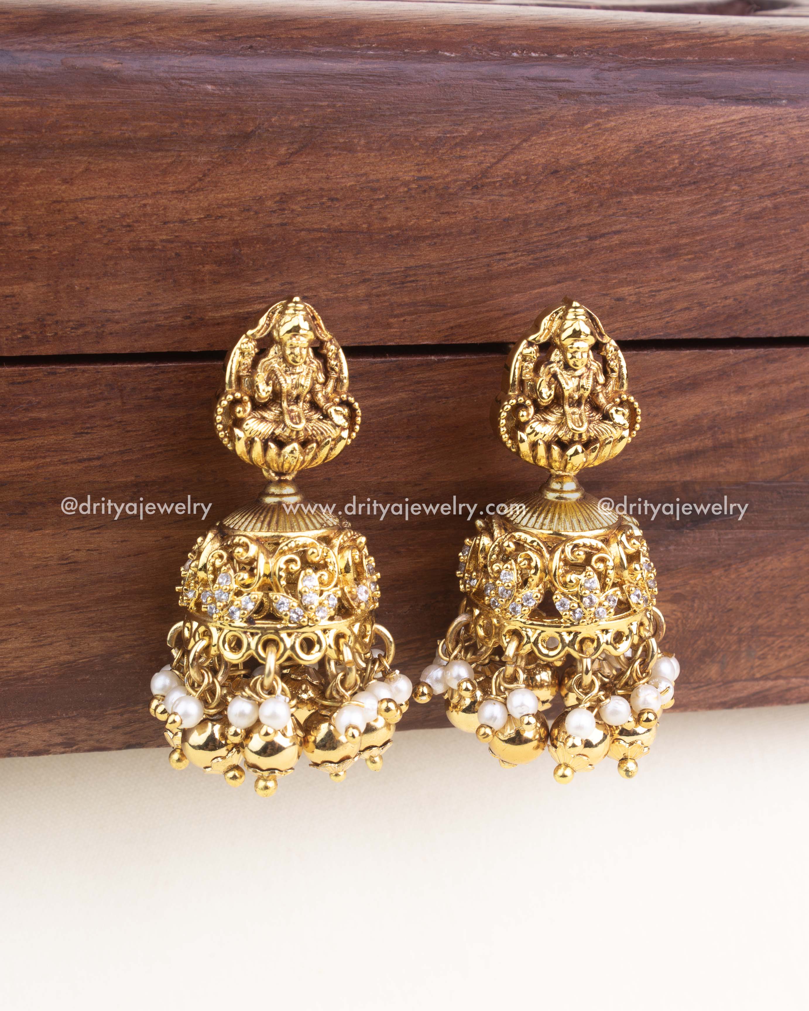 Pair of antique gold jhumka earrings featuring Lakshmi carving, AD accents, and pearl drops