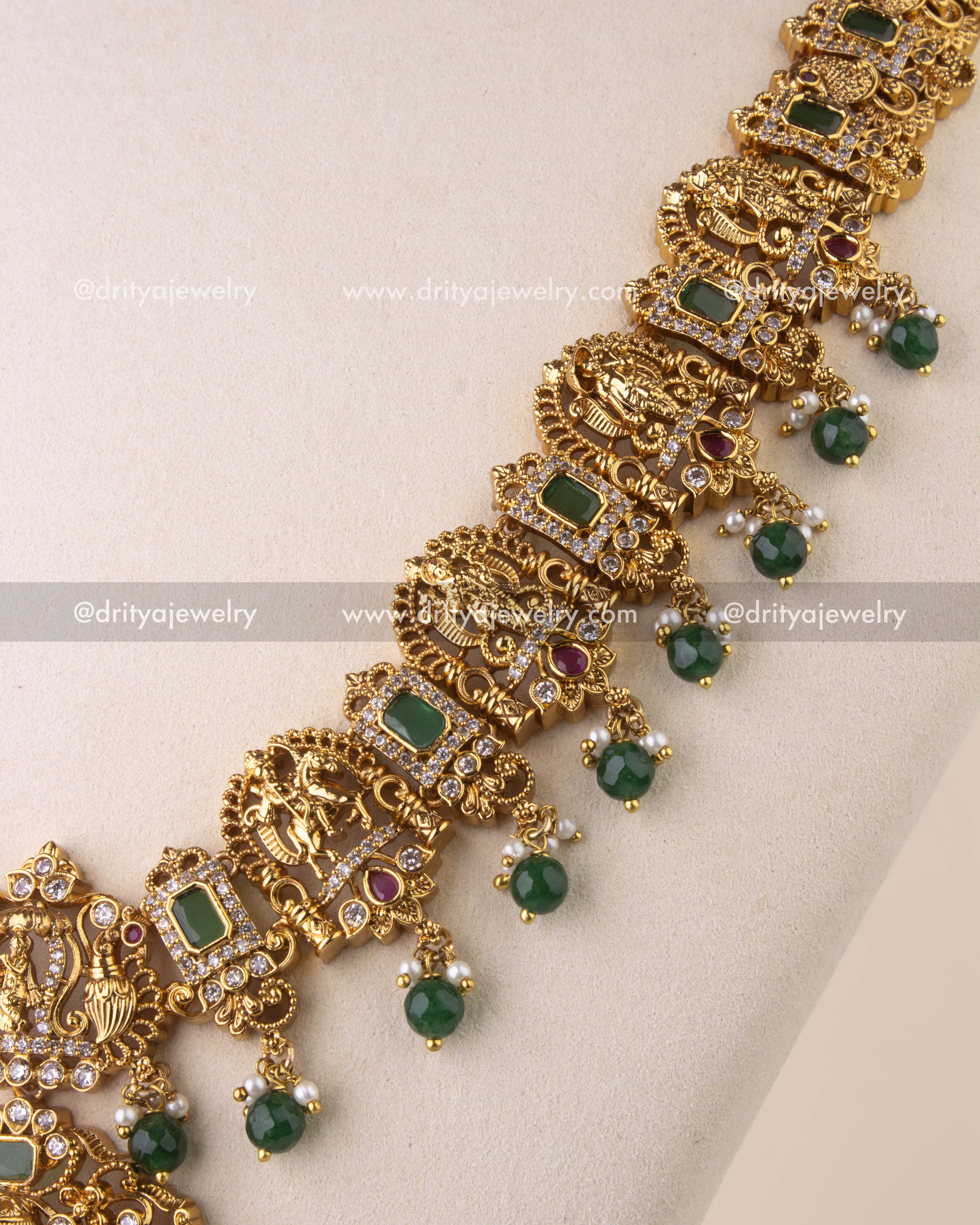Intricate temple chain with goddess and peacock motifs, ruby embellishments, and emerald rectangular stones.