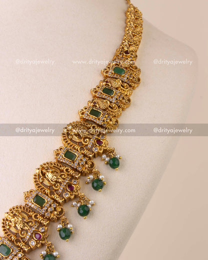 Intricate temple chain with goddess and peacock motifs, ruby embellishments, and emerald rectangular stones.