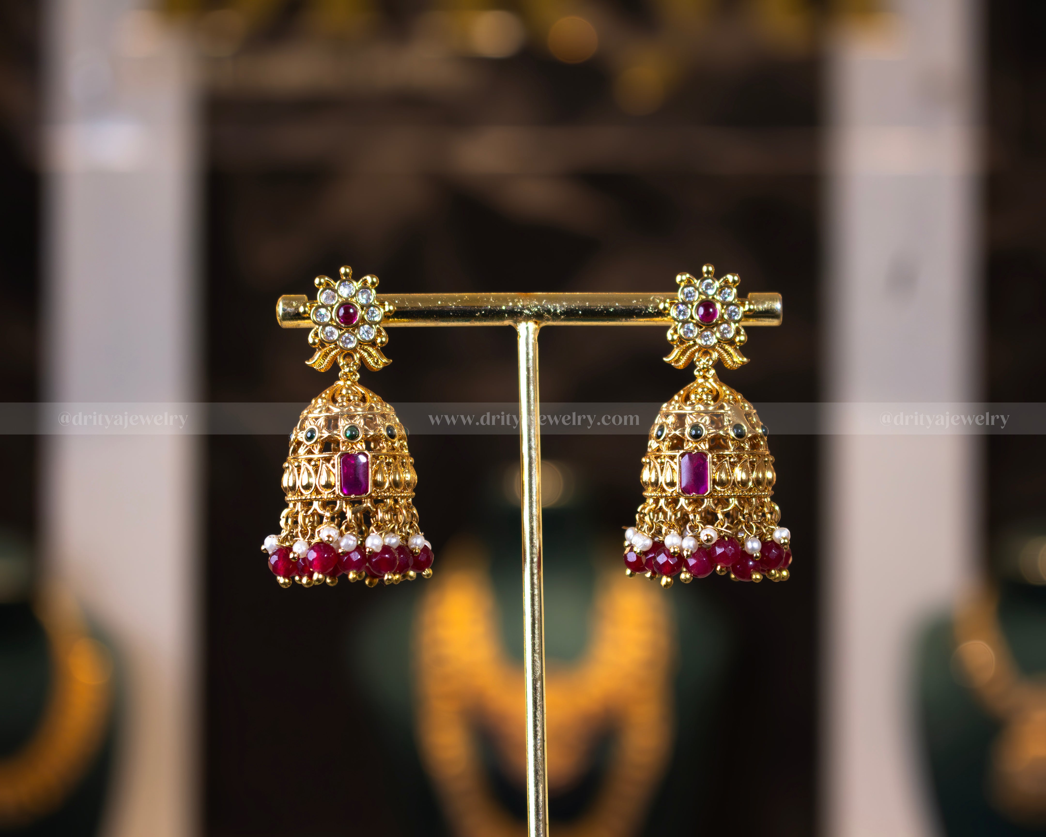 Close-up of Dritya Jewelry's temple-style jhumka earrings with red beads, featuring intricate gold-plated designs and stone embellishments.