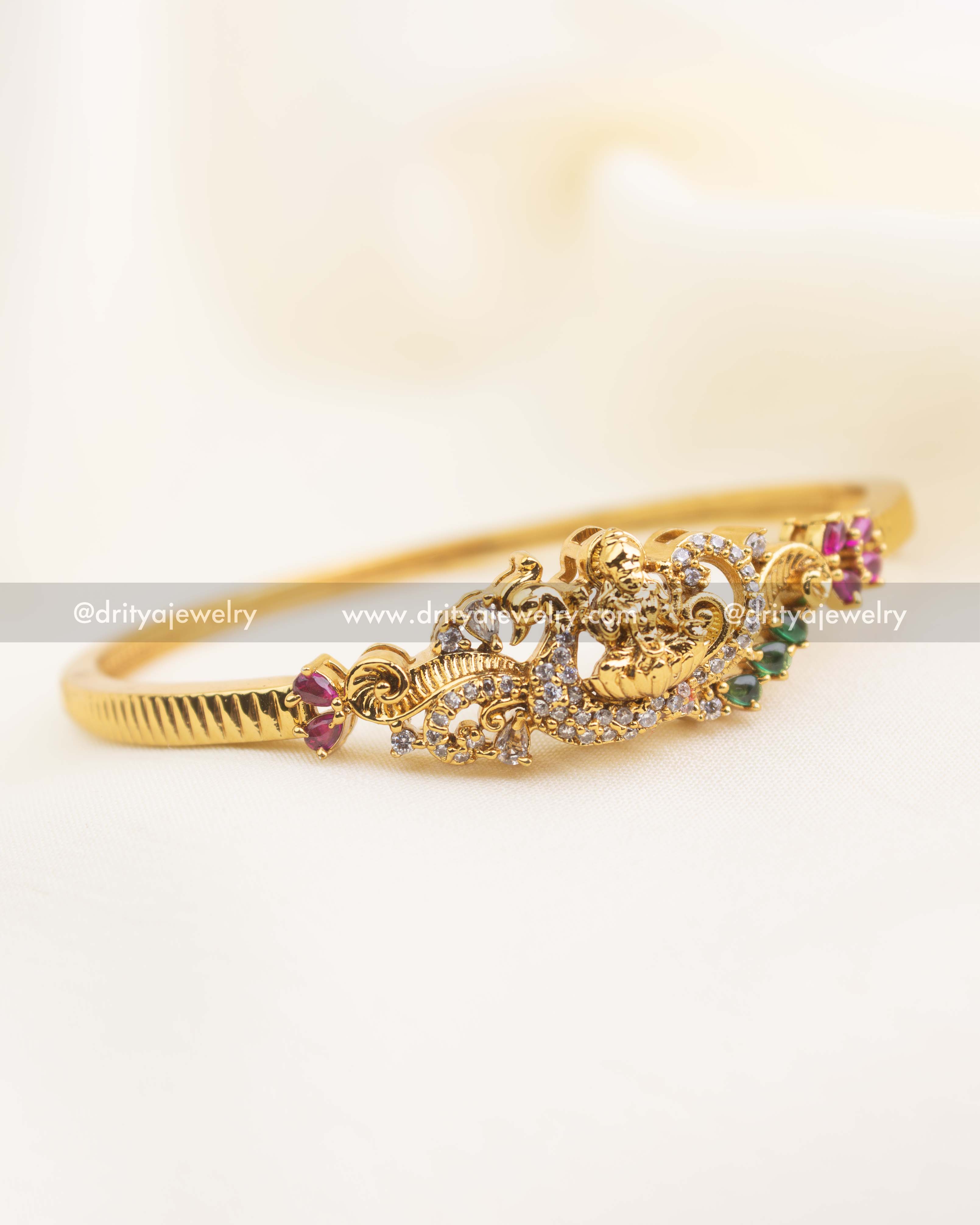 Gold-plated temple kada with ruby and emerald stones in openable design displayed on fabric.